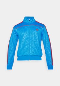 Zils sporta žakets ar rāvējslēdzēju, kas izgatavots no teksturēta materiāla, ar sarkanām akcentu svītrām uz pleciem un mazu logotipu kreisajā krūšu da�ļā.