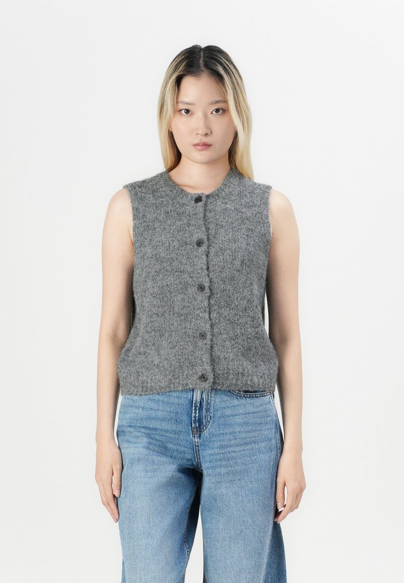 Gilet grigio lavorato a maglia senza maniche con scollatura arrotondata e chiusura a bottoni. Tessuto morbido, aderente in vita, abbinato a jeans azzurri chiari.