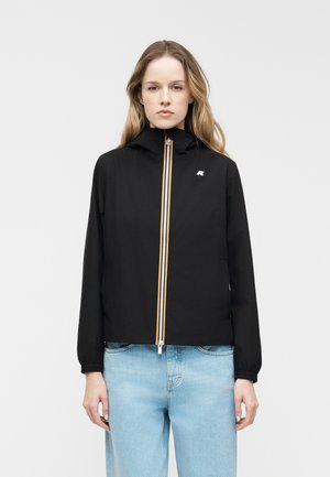 Kvinde iført sort lynlåstrøje med hætte og lyseblå jeans, stående mod en ensfarvet lys baggrund, set forfra.