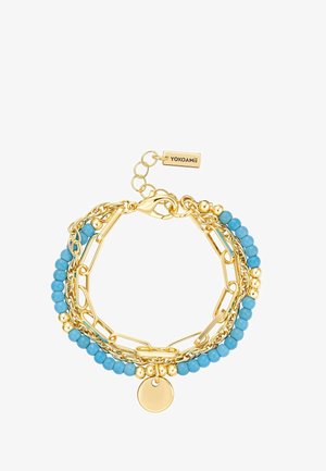 Goldfarbene Mehrfachketten-Armband mit blauen Perlen, einem runden goldenen Anhänger, einem Karabinerverschluss und einem YOKOAMii-Tag.