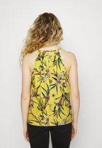 Top halter floral em tecido amarelo com estampado botânico, apresentando um detalhe de keyhole nas costas e um corte descontraído. Combinado com jeans pretos.