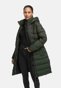 Vrouw in een lange donkergroene gewatteerde jas met capuchon, over een zwarte top en zwarte jeans, poseert met één hand in de zak.
