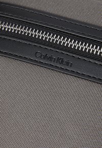 Borsa di tessuto grigia caratterizzata da una cerniera in pelle nera liscia, hardware metallico argentato e logo "Calvin Klein" stampato sull'accento in pelle.