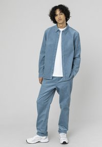 Chemise en velours côtelé bleu clair avec des boutons, assortie à un pantalon en velours côtelé de la même couleur. Un t-shirt blanc en dessous et des baskets blanches complètent la tenue.