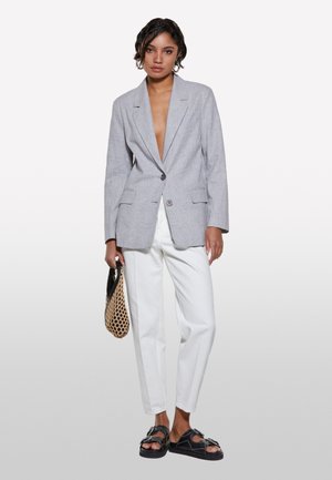 Blazer oversized gris clair avec un col cranté et deux poches avant, associé à un pantalon blanc taille haute et des sandales noires épaisses.