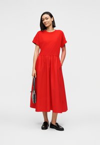 Κόκκινο βαμβακερό φόρεμα μέσης μήκους με κοντά μανίκια και ελαστική μέση. Το μοντέλο κρατά μια μικρή τσάντα χειρός και φορά μαύρα μοκασίνια. Απλός σχεδιασμός, ομαλή υφή.