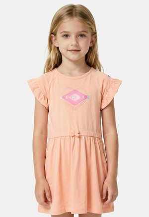 Jeune fille aux longs cheveux blonds portant une robe pêche avec des manches volantées et un motif en forme de cœur sur la poitrine, debout devant un fond uni.