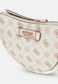 Hvid halvmåneformet Guess-pung med lysebrunt monogrammønster og guldfarvet lynlås samt logo-plakette med teksten "GUESS EST 1981."