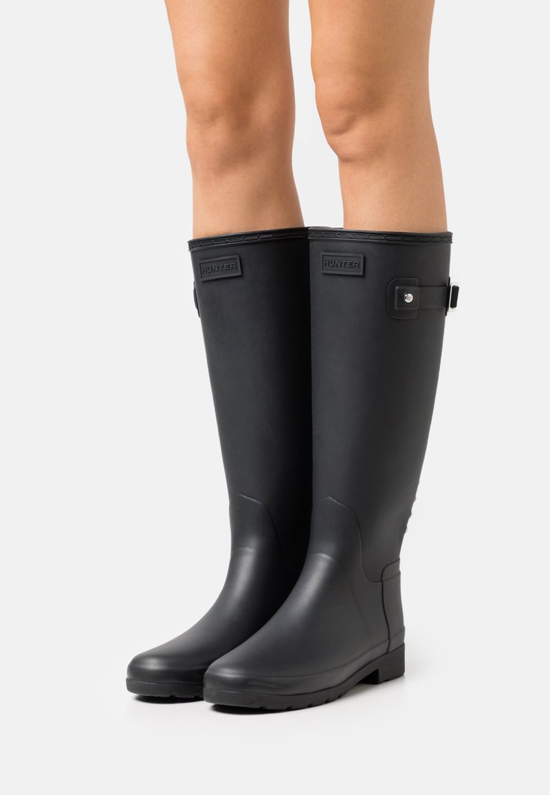 Jambes portant des bottes de pluie en caoutchouc noires montantes jusqu'aux genoux avec une boucle latérale et le logo Hunter sur un fond clair.