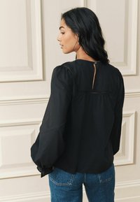 Melna blūze ar puffām piedurknēm, teksturētiem akcentiem un atslēgas caurumu aizmugurē. Saskaņota ar ziliem džinsiem. Vienkāršs un elegants dizains.