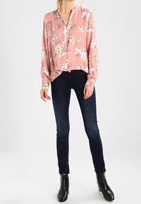 Ljusrosa blus med lång ärm, v-ringning och blommönster i vitt och gult, parat med mörkblå skinny jeans och svarta ankelkängor.