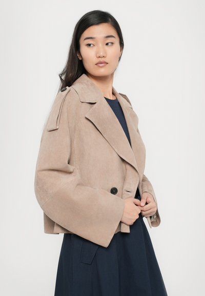 Veste coupée en daim beige avec des manches larges, design double boutonnage, col cranté et boutons noirs, portée sur un haut bleu.