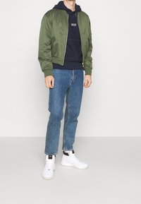 Veste bomber vert olive avec des poignets côtelés, accompagnée d'un sweat à capuche bleu marine avec du texte blanc, d'un jean bleu et de baskets blanches.
