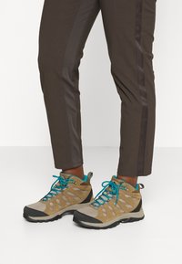 Hnědé turistické kalhoty s bočními pruhy, doplněné lehkými béžovými a šedými turistickými botami s tyrkysovými tkaničkami a síťovinovými panely.