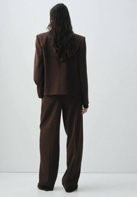 Blazer oversize marron et pantalon large, avec une texture lisse et un design ajusté. L'ensemble a une coupe décontractée avec un minimum de détails.