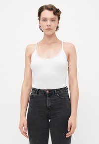 Jeune femme aux cheveux courts portant un débardeur en dentelle blanche et un jean noir taille haute, se tenant devant un fond clair.