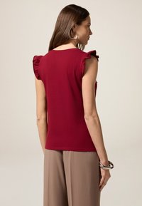 Burgunderfarbene ärmellose Bluse mit Rüschendetails an den Schultern, aus weichem Stoff gefertigt. Kombiniert mit hellbraunen, weiten Hosen und einem silbernen Armreif.