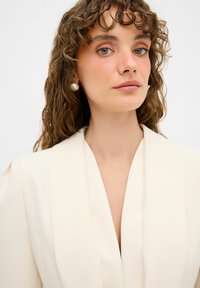 Blazer bianco sporco con profonda scollatura a V, realizzato in tessuto morbido. Presenta spalline morbide e orecchini rotondi perle.