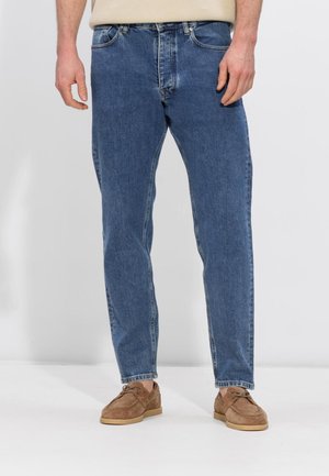 Mann iført blå denimjeans med rette ben og brune snøresko i semsket skinn, stående foran en enkel lys bakgrunn.