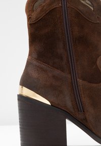 Hnědé semišové kotníkové boty se zipem na boku, s podpatkem se zlatým akcentem a strukturovaným povrchem. Hladký základ podpatku vypadá jako dřevo.