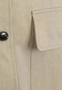Beige overhemd van gestructureerde stof, met een voorzak en een donkere knoop bij de halslijn, die subtiele stikseldetails laat zien.