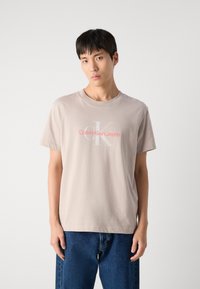 T-shirt beige clair à manches courtes et encolure ronde, arborant un logo rose avec l'inscription "Calvin Klein Jeans" et un design en K gris.
