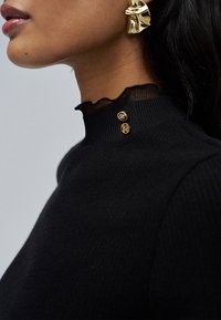 Woman wearing black ribbed high-neck sweater with ruffled sheer trim and two gold buttons, plus large abstract gold earring.