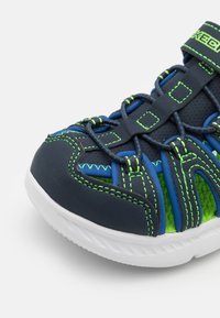 Zapatilla deportiva en color azul marino y verde brillante con un diseño flexible, parte superior de malla, detalles texturizados y sistema de cordones elásticos. Suela blanca.