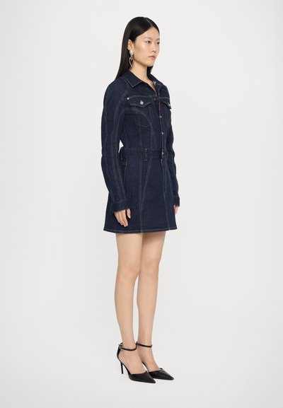 Denim dress in dark blue with long sleeves, belt at the waist, front pockets, and contrasting stitching. Worn with black pointed heels.