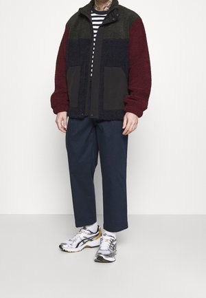 Chaqueta de forro polar en verde, burdeos y negro, con un cuello alto, dos bolsillos frontales y un corte relajado. Combinada con pantalones azul marino y zapatillas plateadas.