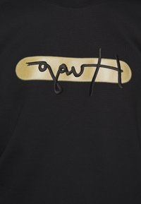 Camiseta de tela negra con un diseño ovalado dorado con texto manuscrito en negro. La textura parece suave con colores contrastantes y delicados.