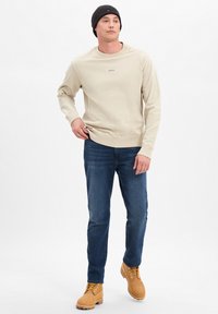 Beżowy sweter z okrągłym dekoltem i subtelnym logo; zestawiony z ciemnoniebieskimi dżinsami i brązowymi sznurowanymi butami. Czarna dzianinowa czapka dopełnia stylizację.