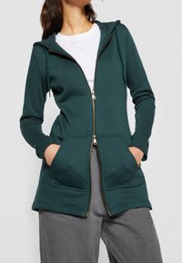 Felpa verde acqua con zip frontale, dotata di due tasche anteriori, tessuto testurizzato, cordini regolabili sul cappuccio, abbinata a pantaloni grigi.