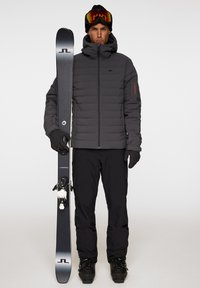 Skiutrustning inkluderar en svart pufferjacka, svarta handskar och byxor. Personen håller en grå skida med vita bindningar.