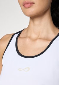 Biely športový tank top s čiernymi prvkami, ktorý má na prednej strane zlaté nekonečné logo. Hladká textúra a okrúhly výstrih.