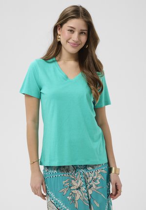 Mlada žena s dugom smeđom kosom, odjevena u tirkiznu majicu s V-izrezom i suknju s cvjetnim uzorkom, smiješi se, nosi zlatne naušnice u obliku obruča i narukvice.