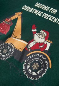 Zielony materiał z kreskówkowym obrazkiem Świętego Mikołaja na żółtym koparko-ładowarce, machającym ręką. Tekst powyżej brzmi „KOPIEMY PREZENTY NA ŚWIĘTA.”