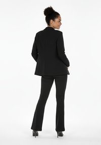 Czarny garnitur szyty na miarę z dopasowaną marynarką i rozszerzanymi spodniami. Gładka tkanina, klasyczne klapy i czysta sylwetka. Całość dopełniają buty na obcasie.