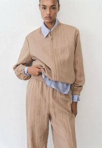 Lagani svijetlosmeđi zip-up jakna s elastičnim manžetama, složena preko svijetloplave košulje s tankim prugama, u kombinaciji s odgovarajućim hlačama.