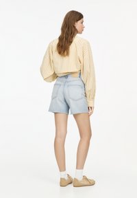 Kremowa, żeberkowa koszula z długim rękawem połączona z wyblakłymi niebieskimi szortami dżinsowymi. Model nosi beżowe buty i białe skarpetki, pokazany z tyłu.