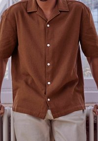 Koszula na guziki z teksturowanej brązowej tkaniny, krótkie rękawy, klasyczny kołnierz, białe guziki, noszona z jasnymi spodniami.