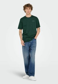 Mörkgrön t-shirt i bomull med korta ärmar, rund hals och liten vit logotyp. Tillsammans med ljusblå vida jeans och vita sneakers.