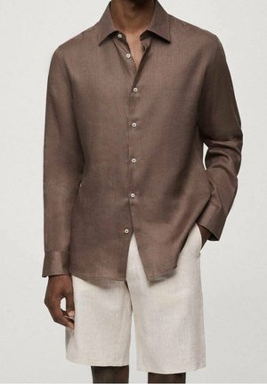 Brązowa koszula na długi rękaw zapinana na guziki, zestawiona z jasnobeżowymi spodniami krótkimi do kolan, jedna ręka w kieszeni, na jednolitym tle.