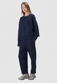 Granatowa bluza i pasujące dresy wykonane z miękkiej tkaniny; luźny krój z prążkowanymi ściągaczami i bocznymi kieszeniami; noszone z szarymi sneakersami.