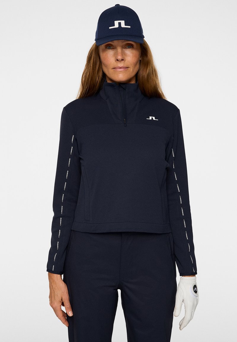 Pullover navy a zip corto realizzato in tessuto morbido, con logo bianco e dettagli a contrasto sulle maniche; abbinato a pantaloni navy coordinati e guanti bianchi.
