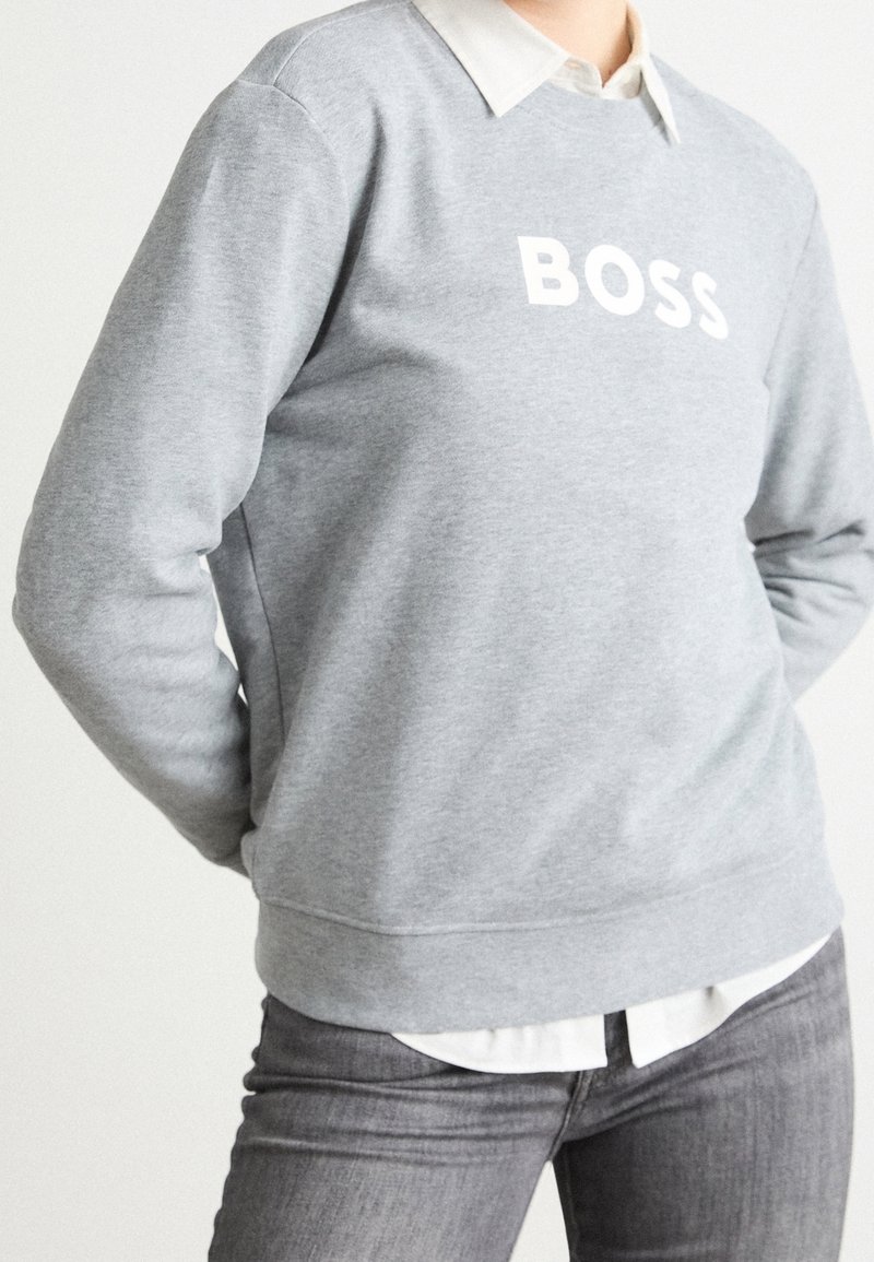 Personne portant un sweat-shirt gris avec le texte "BOSS", une chemise blanche à col en dessous, et un jean gris avec les mains dans le dos.