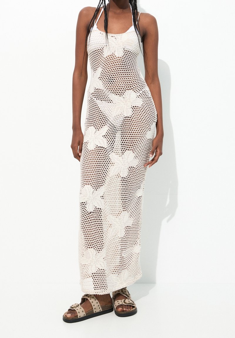 Femme portant une robe maxi en crochet blanche à motifs floraux, bretelles fines et sandales plates beiges cloutées.