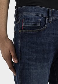 Jeans en denim bleu avec une coupe slim, dotés d'une poche avant avec des coutures rouges, d'un léger délavage et d'une finition texturée.