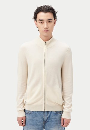 Junger Mann mit lockigem dunklem Haar, trägt eine cremefarbene Strickjacke mit Reißverschluss und blaue Jeans, steht vor einem schlichten hellen Hintergrund.