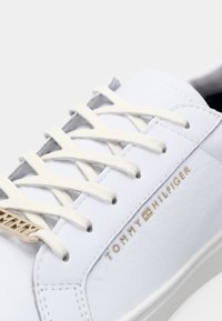 Białe skórzane sneakersy z płaskimi białymi sznurowadłami, złotym logo "TOMMY HILFIGER" z boku oraz gładkim, czystym designem.